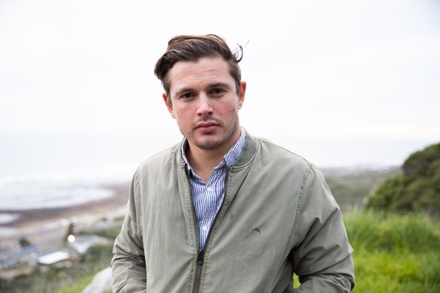 Young man standing in countryside near the sea, wearing casual jacket and shirt, looking at camera. Ideal for use in lifestyle blogs, travel websites, outdoor adventure promotions, and personal development articles.