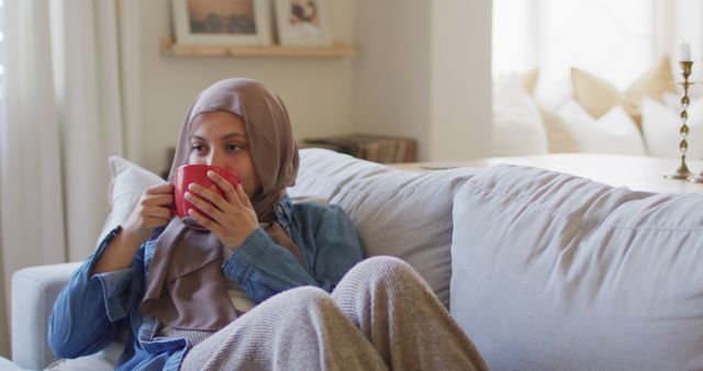 Image of thoughtful biracial woman in hijab drinking coffee relaxing on sofa in living room at home. Happiness, relaxation, inclusivity and domestic life.