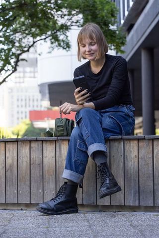 Young woman in her 20s sitting on urban bench holding smartphone and smiling