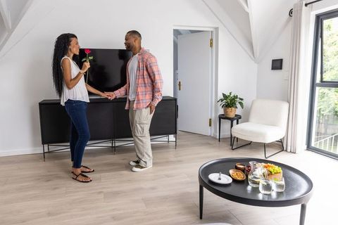 Couple exchanging rose while holding hands in modern sunlit loft living room, cozy decor