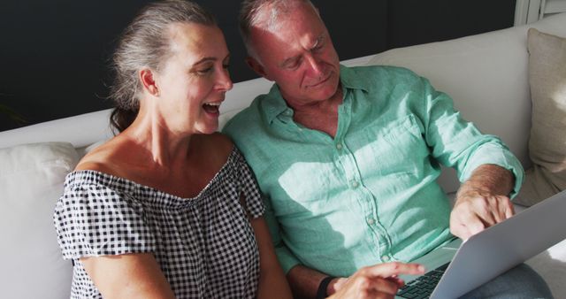 Caucasian senior couple sitting on sofa talking in the sun using laptop. Staying at home in isolation during quarantine lockdown.