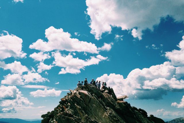 Group of hikers standing on rocky mountain peak under clear sky with fluffy clouds. Represents teamwork, accomplishment, and outdoor adventure. Ideal for use in travel blogs, adventure magazines, and outdoor gear advertisements.