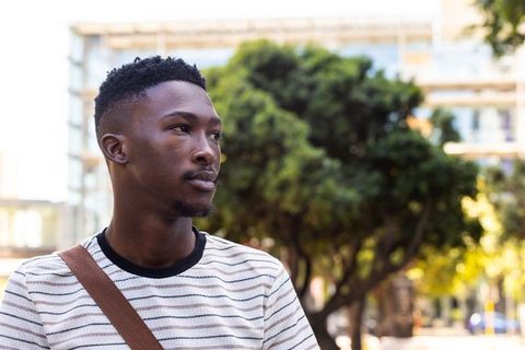African man standing on urban sidewalk wearing striped shirt and crossbody bag looking thoughtful