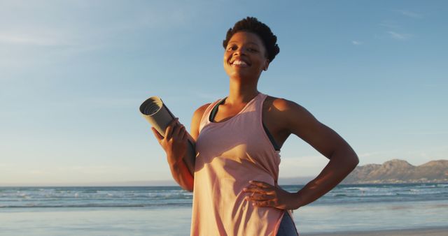 Portrait of african american woman holding yoga mat at the beach smiling. healthy active lifestyle, outdoor fitness and wellbeing.