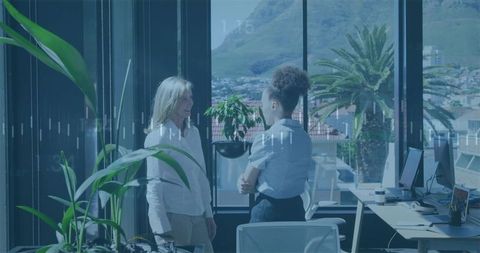 Two coworkers talking in modern glass office with city skyline and greenery