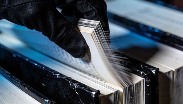 Gloved hand flipping archival ledger pages under blue light revealing worn spines and pale edges