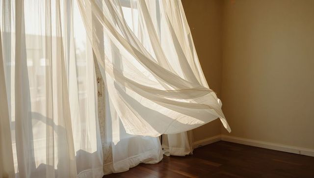 Sunlit sheer curtains billowing over warm hardwood floor near glass door