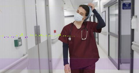 Nurse adjusting hair cover in hospital corridor wearing maroon scrubs and surgical mask