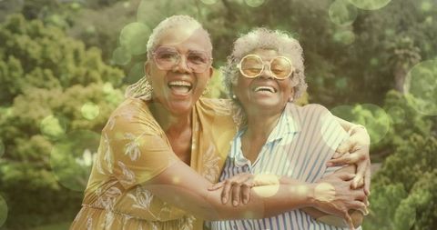 Laughing joyful senior women embracing in sunlit garden wearing vintage sunglasses