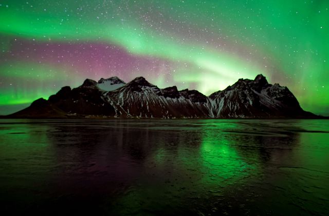 Vivid Northern Lights illuminating snow-capped mountains and reflecting in a calm body of water in Iceland. The green and purple hues create a mesmerizing night sky display. Ideal for use in travel promotions, nature documentaries, and inspirational wallpapers showcasing natural beauty.