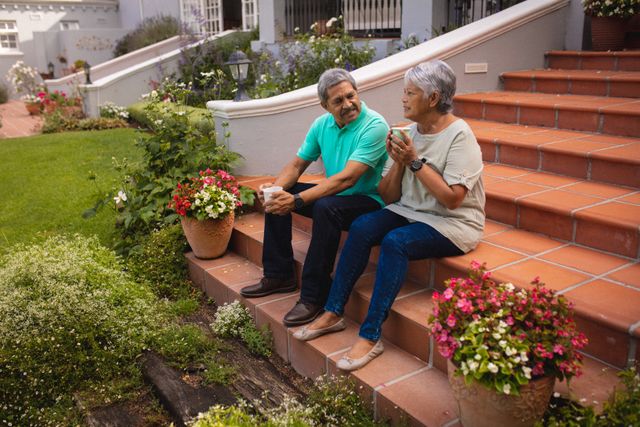 Biracial senior couple with coffee cups talking while sitting on staircase against house in yard. drink, love, togetherness, unaltered, lifestyle and retirement concept.