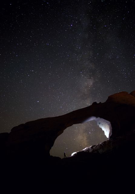 Person standing under natural rock archway silhouetted against star-filled sky. Milky Way galaxy visible. Perfect for themes of stargazing, astronomy, night adventures, natural wonders, and the beauty of dark skies. Ideal for travel, adventure content, and nature or astronomy blogs/websites.