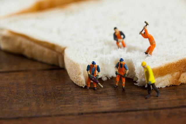 Miniature workers in construction gear are seen working on a slice of bread, creating a humorous and creative scene. This image can be used for concepts related to teamwork, creativity, and food. It is ideal for advertising, social media posts, and editorial content that aims to capture attention with a unique and playful twist.