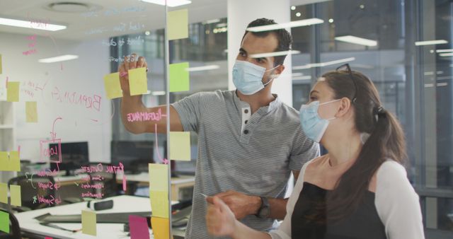 Office workers are brainstorming and collaborating by writing on sticky notes stuck to a glass wall. Both employees are wearing masks, indicating ongoing safety measures due to the pandemic. They seem focused and engaged in planning ideas. Use this image for illustrating teamwork, corporate settings, health precautions in the workplace, or business planning activities.