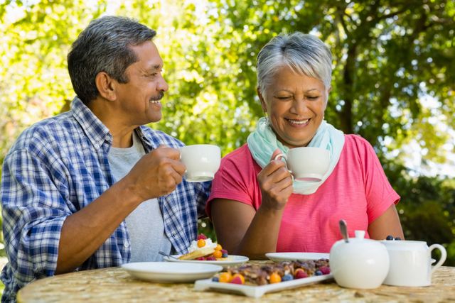Senior couple enjoying tea in a park, smiling and bonding over a relaxing outdoor picnic. Ideal for use in advertisements promoting healthy lifestyles, retirement communities, senior activities, and family bonding moments.