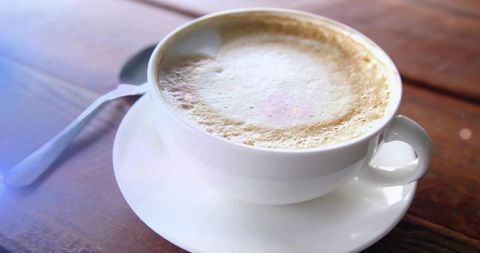Sitting White Cup Showing Frothy Latte on Wooden Table with Spoon and Soft Light Flare