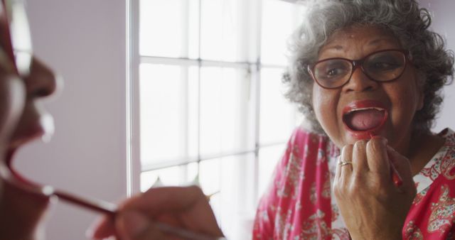 A senior woman is putting on lipstick while looking in the mirror, smiling with eyeglasses on. Image can be used for topics related to beauty routines for older women, promoting self-care and confidence, aging gracefully, makeup tutorials and products for seniors.