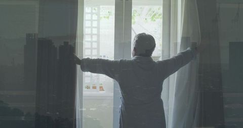 Senior woman opening curtains to morning light at home window