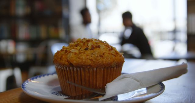 Close-up of a freshly baked muffin placed on a plate with a fork and napkin in cozy café. Ideal for use in blogs, advertisements, and social media posts focused on food, bakery products, coffee shops, and inviting atmospheres. Perfect for highlighting baked goods, breakfast options, or promoting local cafés and bakeries.