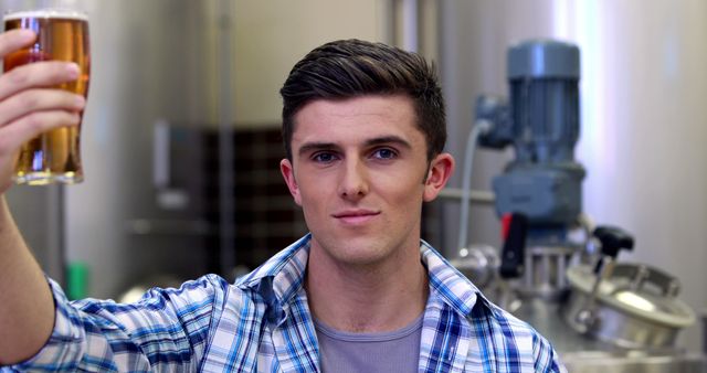 Young man holding up a glass of beer, standing in a brewery with industrial machinery in the background. He appears to be evaluating or inspecting the beer, suggesting themes of quality control or craft brewing process. Useful for representations of brewing industry, craft beer production, and quality assurance.