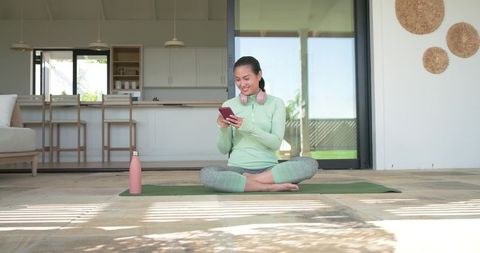 Asian woman sitting cross-legged on patio yoga mat using smartphone with pink water bottle