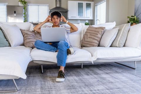 Young asian man working remotely on laptop wearing headphones on white sectional sofa