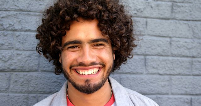 Happy biracial man with brown curly hair standing and smiling by wall on sunny street. City break, summer, travel, vacations and lifestyle, unaltered.