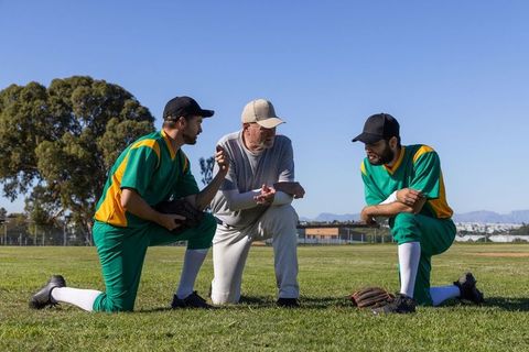 Senior Coach Coaching Two Baseball Players Kneeling on Grass Field Discussing Strategy