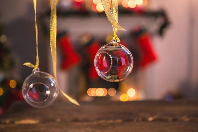 Close-up view of glass Christmas ornaments hanging on golden ribbons in a cozy home setting. The background features a fireplace adorned with stockings and warm lights, creating a festive atmosphere. Ideal for holiday greeting cards, seasonal marketing materials, and home decor inspiration.