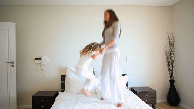 Mother and daughter appear to be having fun together, jumping on a bed in a hotel room with white decor. The room is well-lit and features modern furniture, offering a clean and inviting atmosphere. This cheerful video can be used in marketing materials for family-friendly hotels, travel advertorials, or articles focused on family bonding and activities.