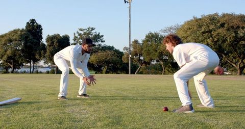 Two cricketers fielding in white kit on sunlit grass with red ball and bat