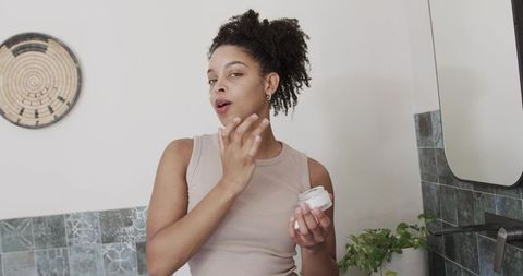 African american woman applying face cream at mirror for minimalist skincare routine