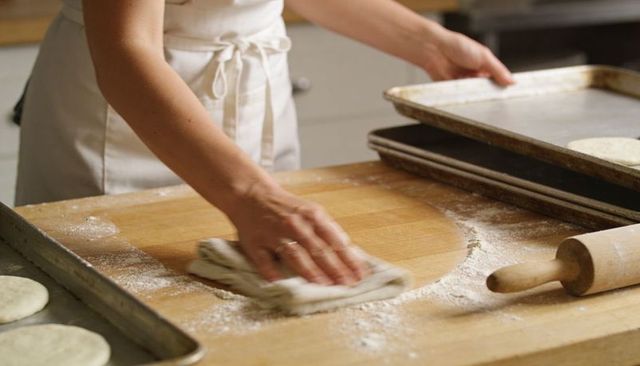 Baker wiping flour from wooden workboard with cloth, rolling pin and sheet pans