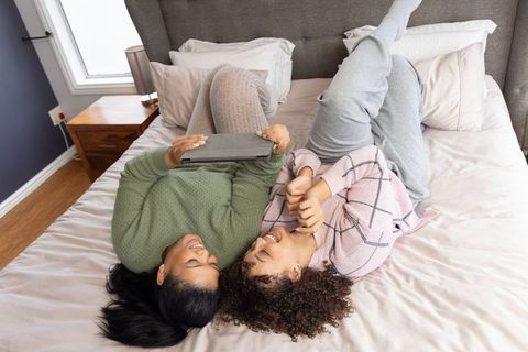 Multicultural friends laughing and using tablet while relaxing on cozy sunlit bed at home