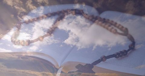 Rosary resting on open bible overlaid with cloud-filled sky double exposure spiritual