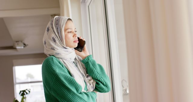 Young Muslim woman in hijab talking on smartphone while looking out window. Ideal for concepts of communication, technology, cultural diversity, modern lifestyle, and thoughtful moments. Can be used in advertisements, blogs about modern communication or cultural topics, and social media posts focusing on daily routines.