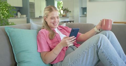 Young woman reclining on sofa smiling while scrolling smartphone and holding coffee mug