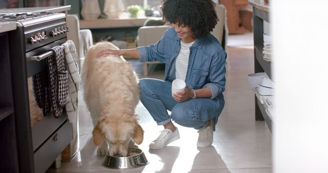 Woman feeding a golden retriever in a cozy home kitchen. She is wearing a casual outfit and smiling warmly at her pet as it eats from a stainless steel bowl. Ideal for use in articles about pet care, canine nutrition, home life with pets, and the human-animal bond.