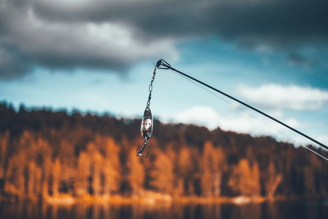 Fishing rod hanging with lure by tranquil lake with autumn forest background. Ideal for content about outdoor activities, fishing enthusiasts, nature exploration, and recreational hobbies.