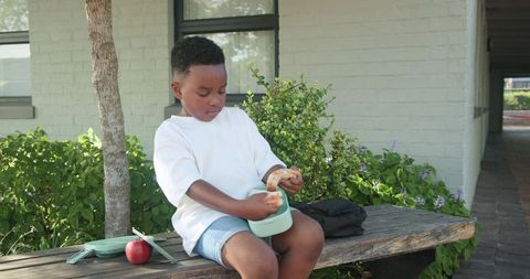 African american boy eating lunch on courtyard bench with backpack and apple during school break