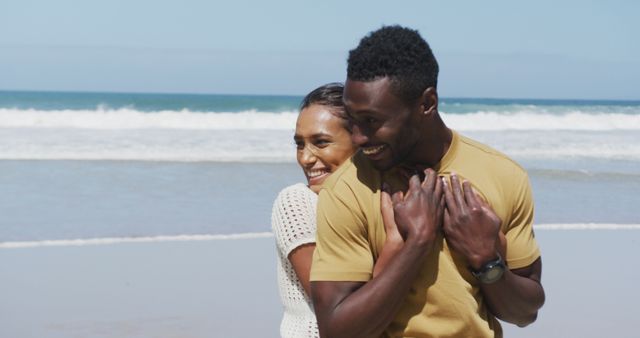 Happy couple embracing and enjoying a sunny day at the beach with ocean waves in the background. Great for themes of love, romance, summer vacations, travel, and joyful moments shared by young adults.