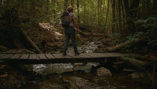 Male hiker pausing on wooden footbridge over creek in dense forest carrying large backpack