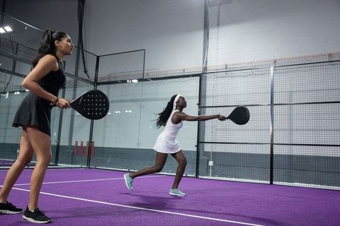 Women playing padel doubles on vibrant purple court lunging to return forehand