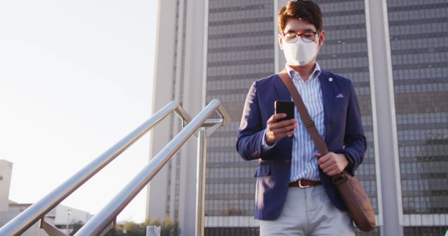 Asian man wearing face mask using smartphone while walking down the stairs at corporate park. health protection and safety during covid-19 pandemic concept