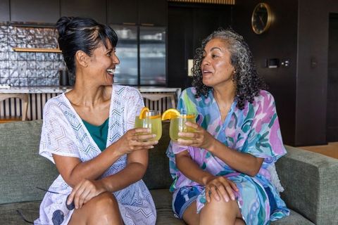 Two african american women toasting citrus cocktails on modern sofa laughing
