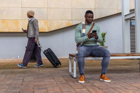 Urban commuters using smartphones while sitting on bench, traveler pulling suitcase