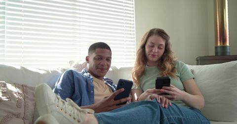 Young couple relaxing on couch using smartphones, sharing moments in bright living room