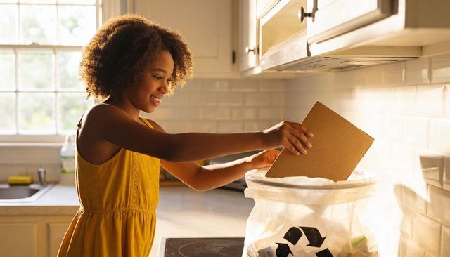 Young girl placing cardboard into clear recycling bag in sunlit kitchen, learning chores
