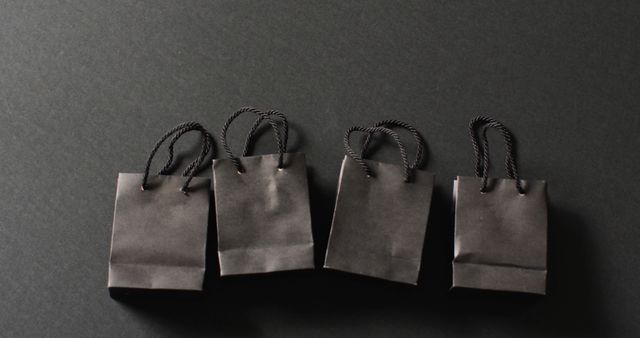 Four black gift bags with black handles in a row on dark grey background with copy space. Luxury treat, present, shopping, black friday sale and retail concept digitally generated image.