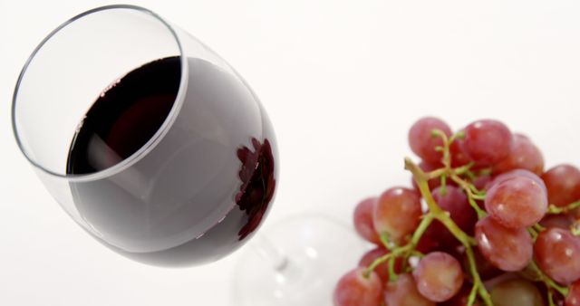 This image shows a glass of red wine next to a bunch of fresh red grapes on a white background. Ideal for use in advertising for wineries, articles about wine health benefits, gourmet or culinary blogs, or promotional material for gourmet dining experiences.